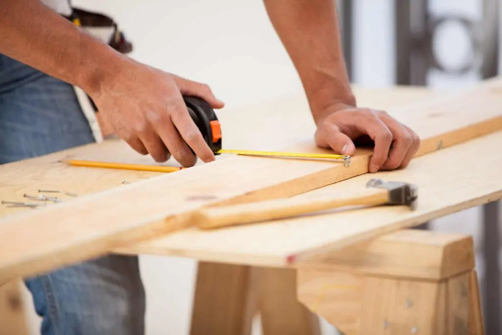 Person measuring wood with a tape measure and pencil on a workbench, surrounded by tools, illustrating home renovation and construction practices.