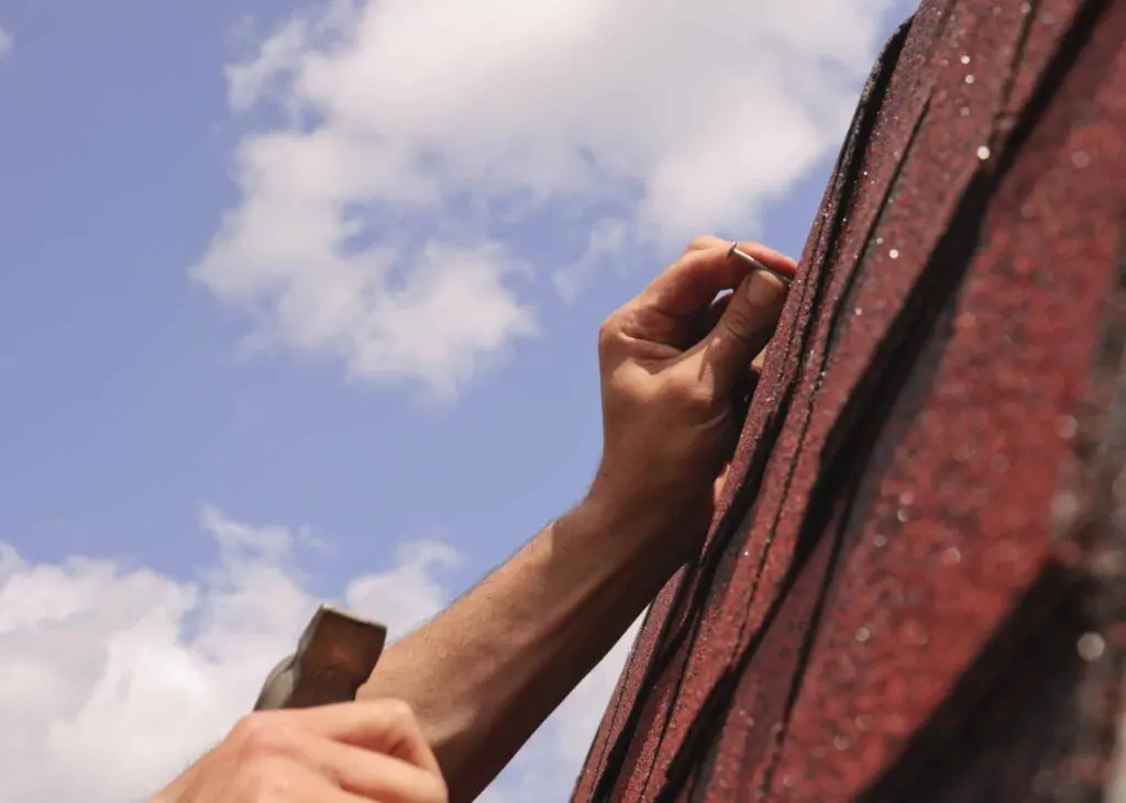 Hand hammering shingles on a roof under a blue sky, illustrating roofing repair and maintenance relevant to insurance coverage for roof replacement.
