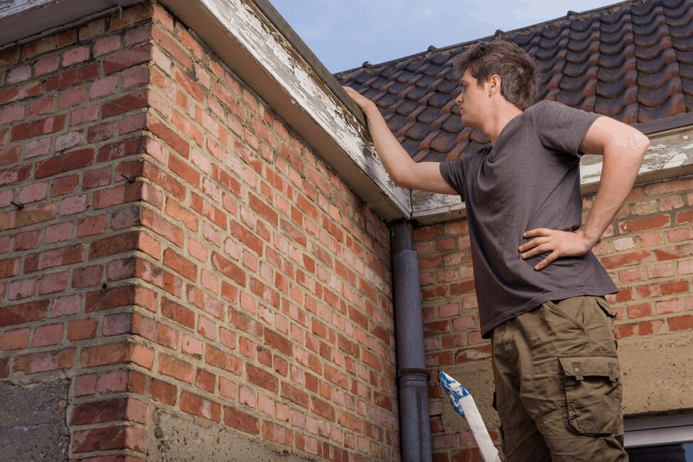 Man inspecting the edge of a roof for potential leaks and damage, with a focus on the flashing and brick wall, emphasizing roof maintenance and inspection techniques.
